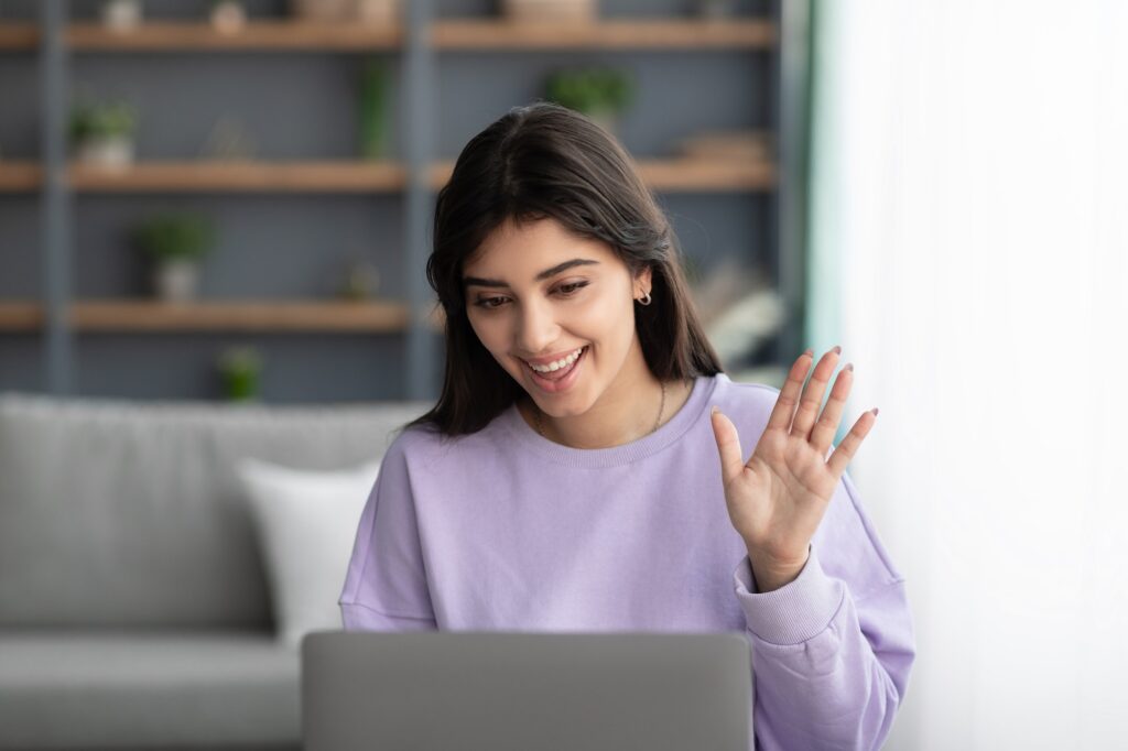 Cheerful woman having videocall using laptop and waving hand